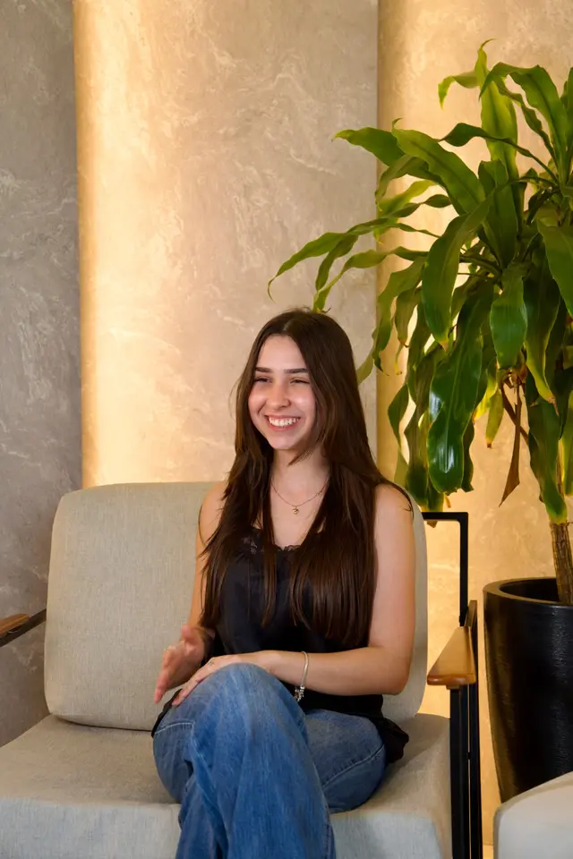 Eduarda, sócia da Solura Studio, de blusa preta e calça jeans, sorrindo sentada em uma poltrona ao lado de uma planta