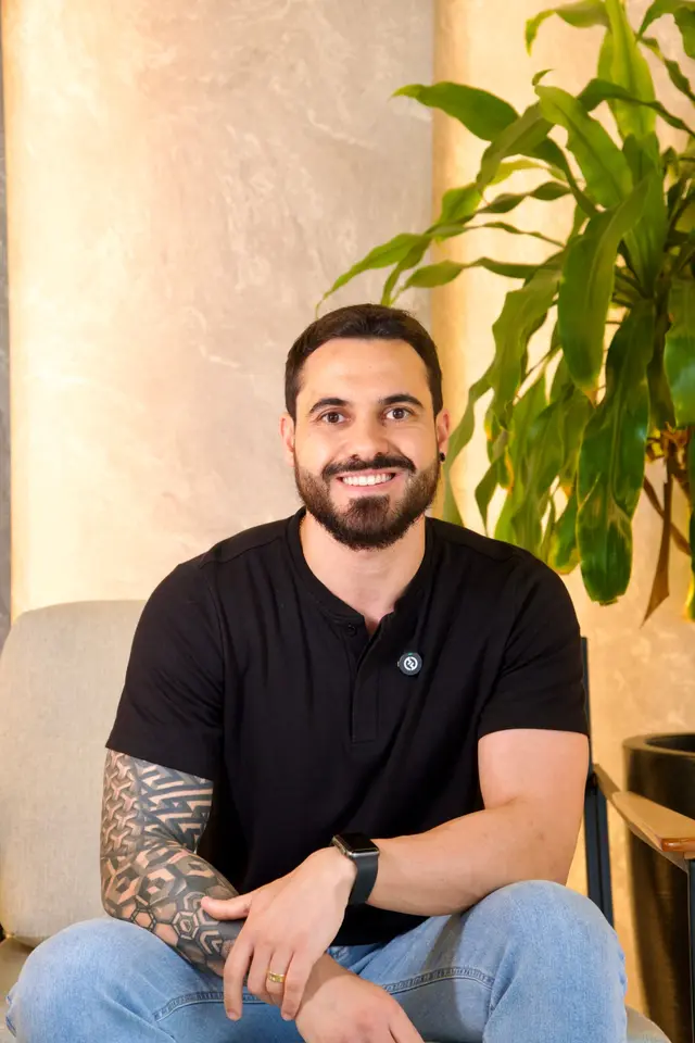 Lucas, sócio da Solura Studio, de camiseta preta, sorrindo sentado em uma poltrona clara diante de uma parede iluminada e de uma planta